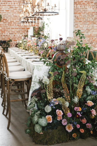 Luxurious wedding reception table setup in an industrial-chic loft with exposed brick walls, exuding elegance and sophistication. The long banquet table is draped in a premium white linen cloth with intricate green botanical embroidery, while lavish flora