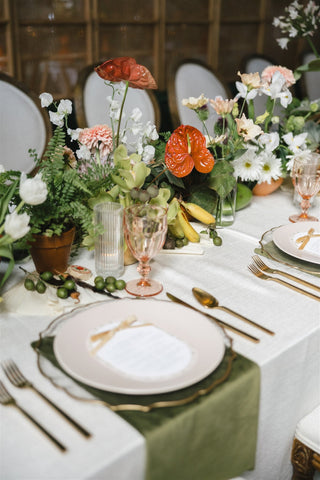 A beautifully arranged dining table featuring elegant plates and glasses, complemented by a floral centerpiece consisting of cut flowers in a vase. The table is adorned with a decorative tablecloth and candles, set against a backdrop that includes plants 